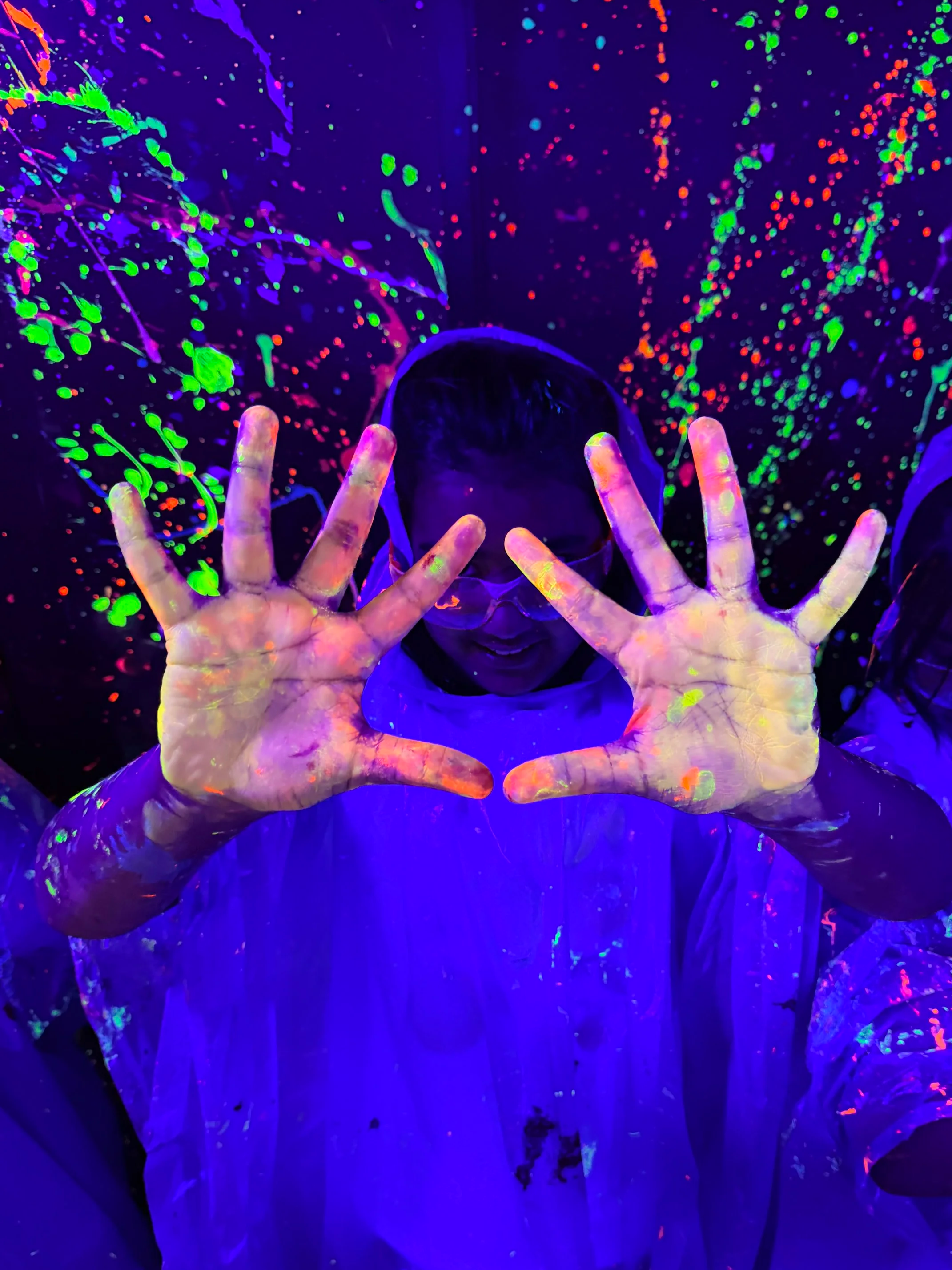 Person with hands raised, covered in glow-in-the-dark paint, smiling and surrounded by neon splatters on dark background.