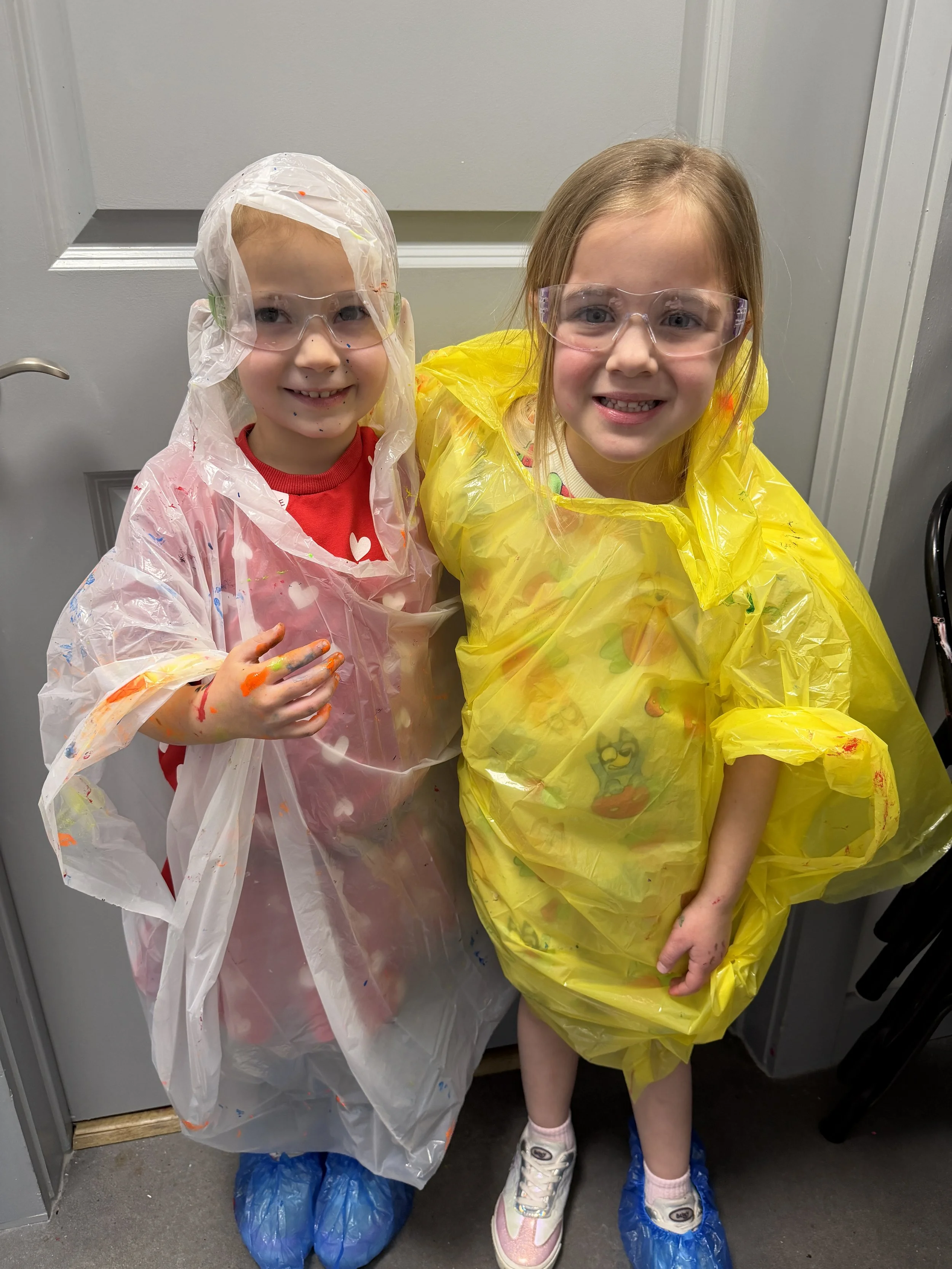 Two young girls wearing colorful rain ponchos and protective glasses, standing inside a building near a door, smiling and showing their painted hands.
