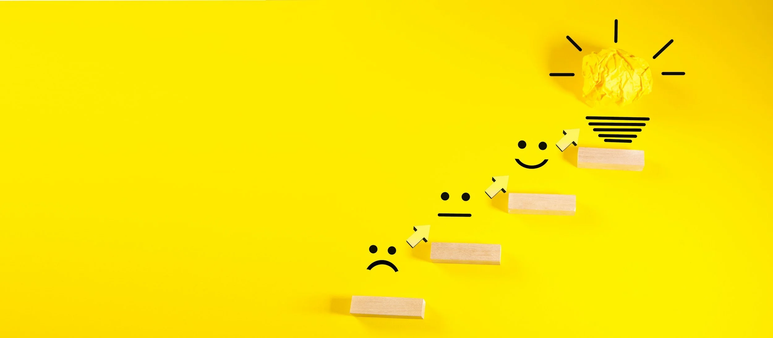 A yellow background with wooden blocks arranged in a staircase pattern, each with a drawn face showing various emotions from sad to happy. A yellow crumpled paper ball representing a light bulb is at the top, with black lines radiating outward, symbolizing an idea or inspiration.