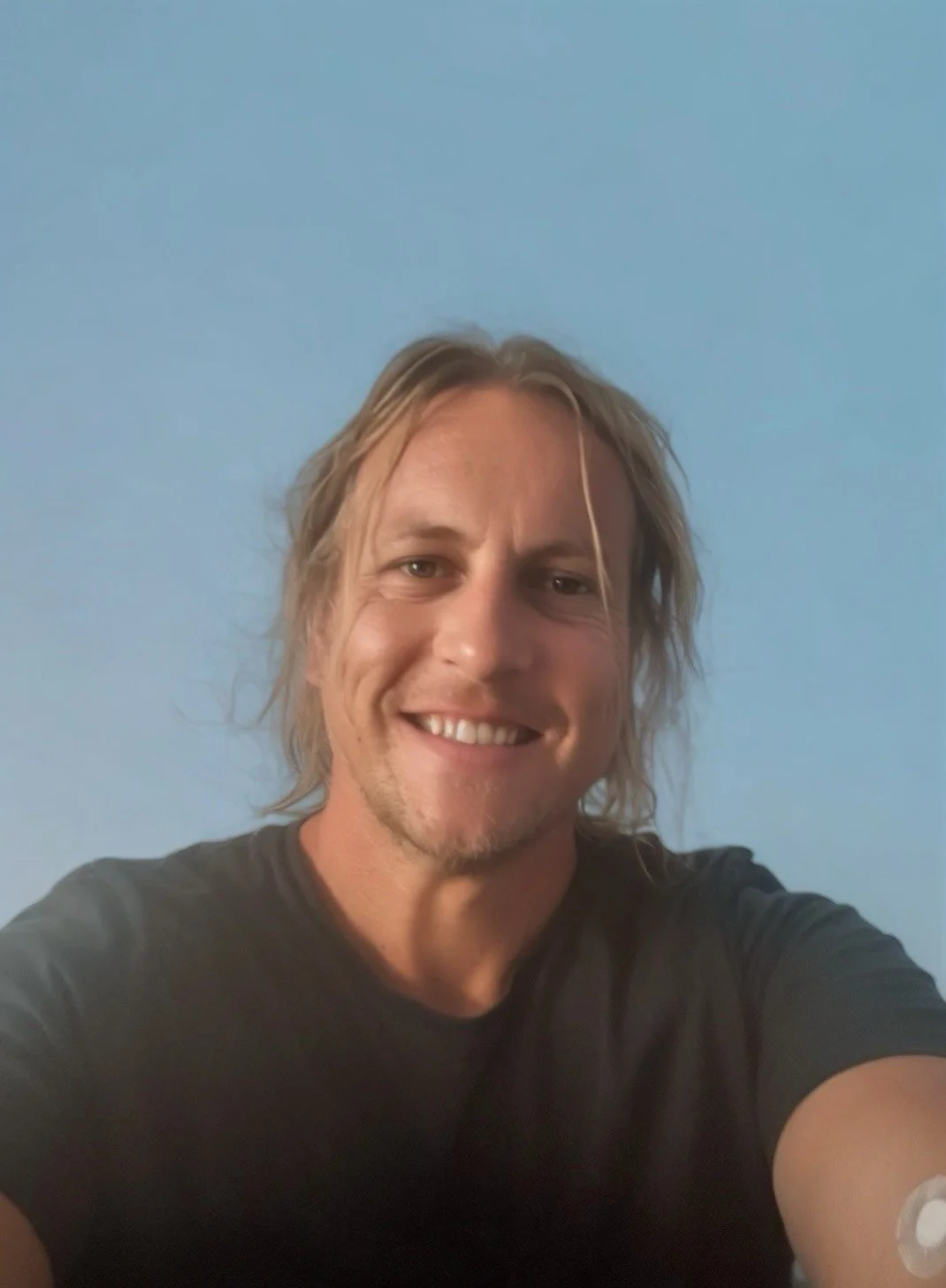 A man with long, light brown hair and a goatee taking a selfie against a plain blue wall, smiling at the camera.