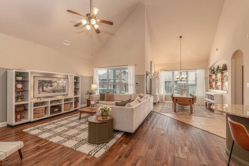 A spacious living room with a vaulted ceiling, ceiling fan, large windows with white curtains, and a cozy seating area with a white sofa. There is a TV on a white bookshelf, a coffee table, and a rug. Adjacent to the living area is a dining space wit