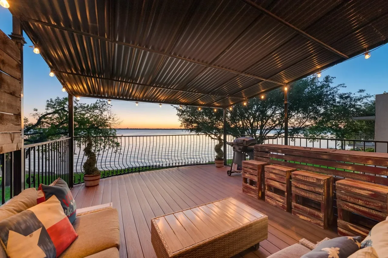 An outdoor patio with wooden flooring and a metal roof, decorated with string lights. It features a sofa with cushions, a wooden coffee table, potted topiary plants, a barbecue grill, and views of trees and a body of water at sunset.