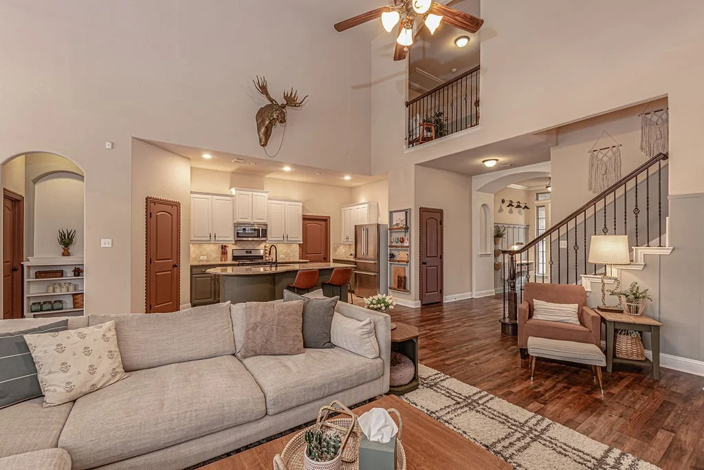 Living room with beige sofa, brown armchair, and wooden side table, open to kitchen with white cabinets, stainless steel appliances, and a vaulted ceiling with a ceiling fan, wood flooring, and decorative wall-mounted moose head