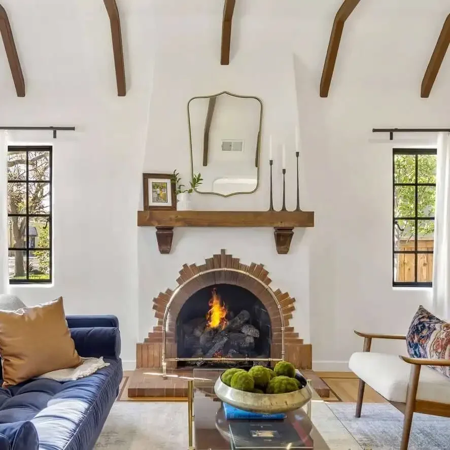 Bright living room with white walls, exposed wooden beams on the ceiling, and large arched windows. Features a white brick fireplace with a mantle decorated with plants, lanterns, and a clock. Contains a white sofa with pillows, checkered armchairs, a wooden coffee table, and a large potted plant.