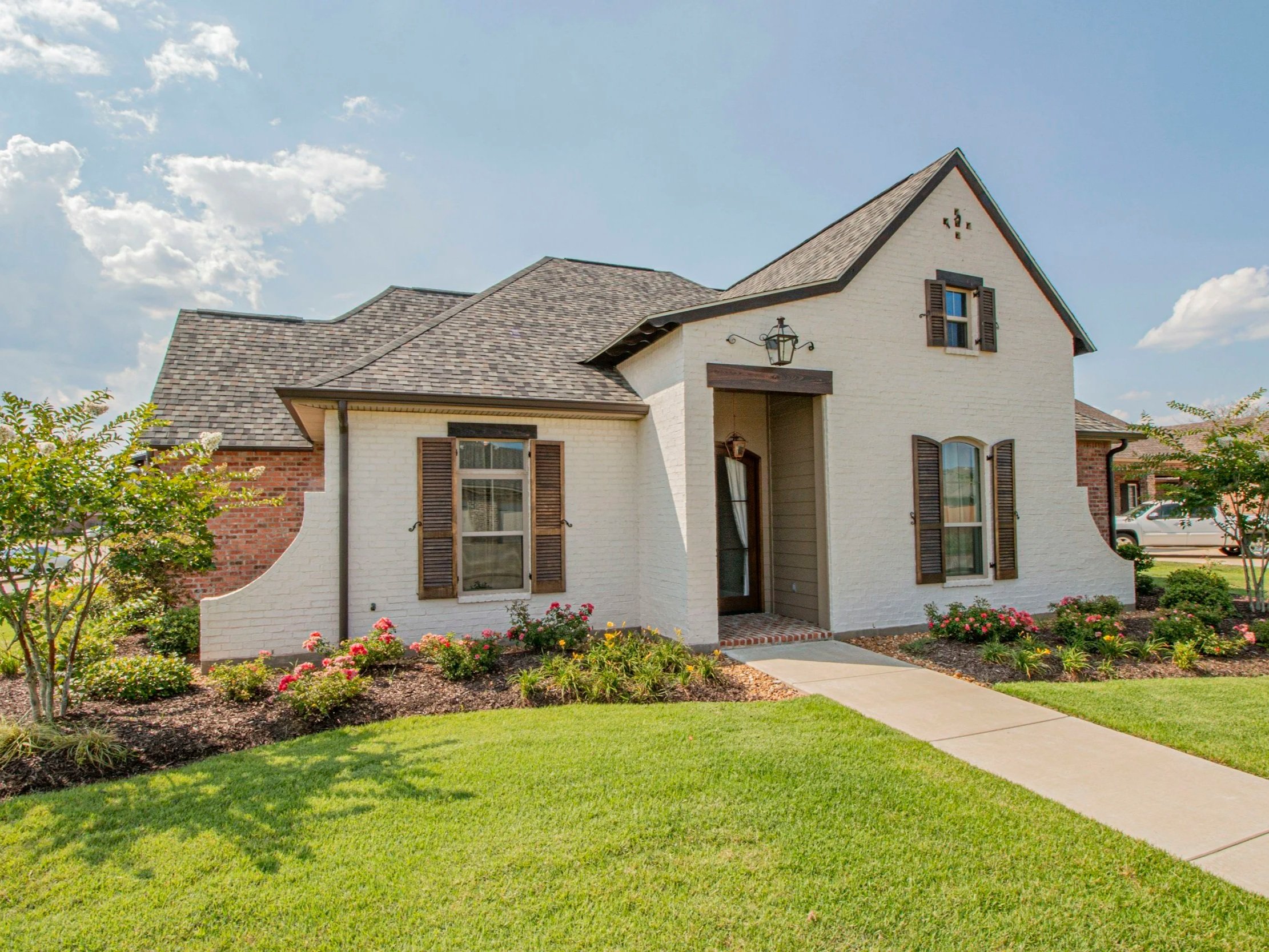 A two-story house with white brick walls, brown shutters on the windows, a brick pathway, a well-maintained lawn, and a garden with bushes and pink flowers, under a partly cloudy sky.