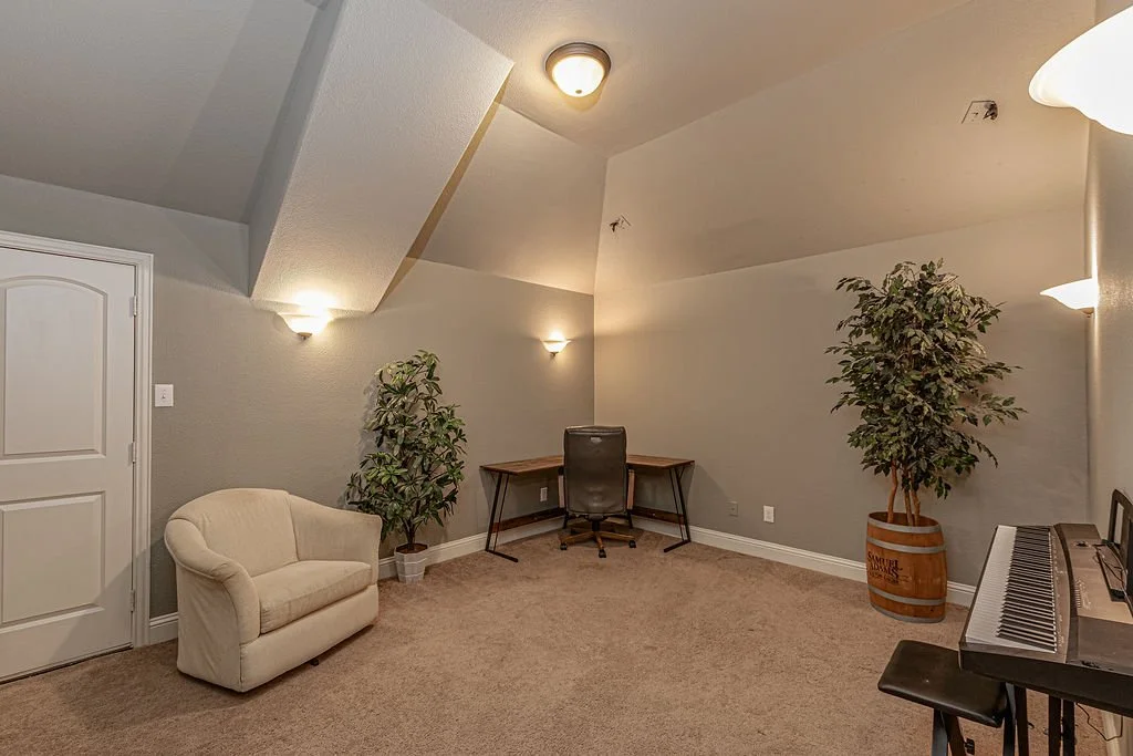 A spacious room with beige carpet, gray walls, and multiple light fixtures. The room contains a small cream-colored sofa, two potted plants, a wooden desk with a black office chair, and a digital piano with a bench. The room has a sloped ceiling and 