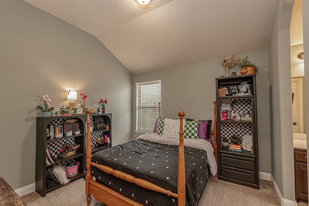 A bedroom with a wooden bed frame, two black bookshelves with checkered backing, and a window with closed blinds.