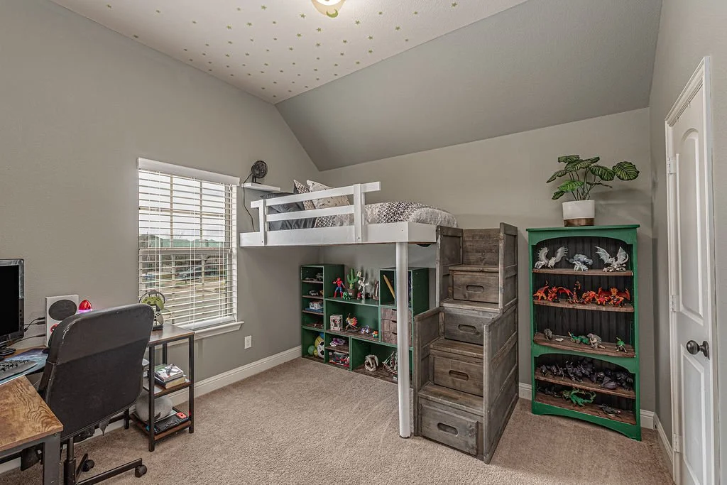 Bedroom with a loft bed, a desk and chair, and green bookshelves filled with toys and figures, beige carpet, window with blinds, ceiling decorated with star and moon decals.