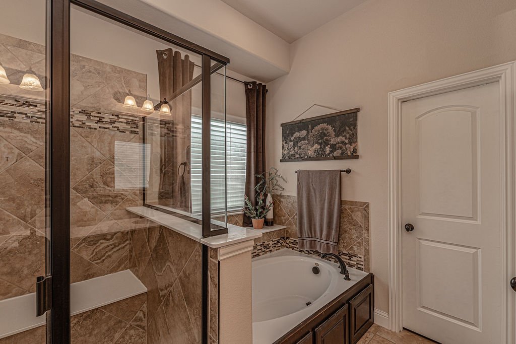 A bathroom with a glass enclosed shower, a bathtub with a wood cabinet, a small window with blinds, a towel rack with a towel, a wall art, and a potted plant on the ledge between the shower and the tub.