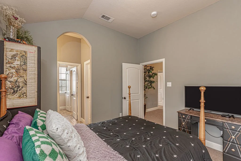 View of a bedroom with gray walls, a bed with black and white bedding, pillows, a TV on a TV stand, and two open doors leading to other rooms.