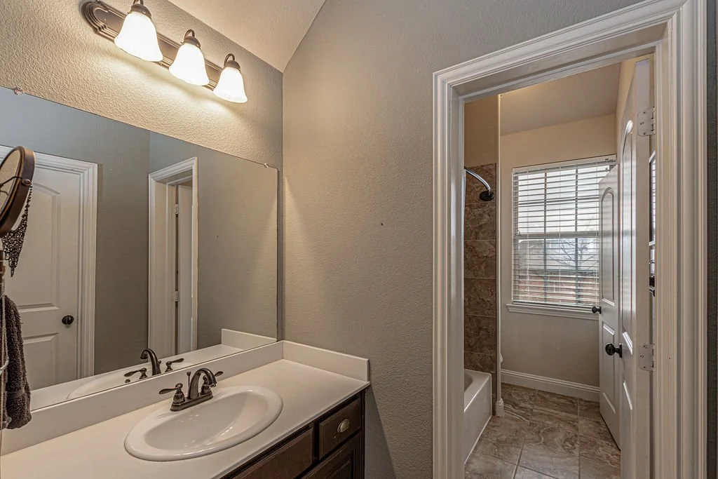 Bathroom with a single sink vanity, large mirror, white countertop, and a three-light fixture above. In the background, there is a shower with tan tile, a bathtub, and a window with blinds.