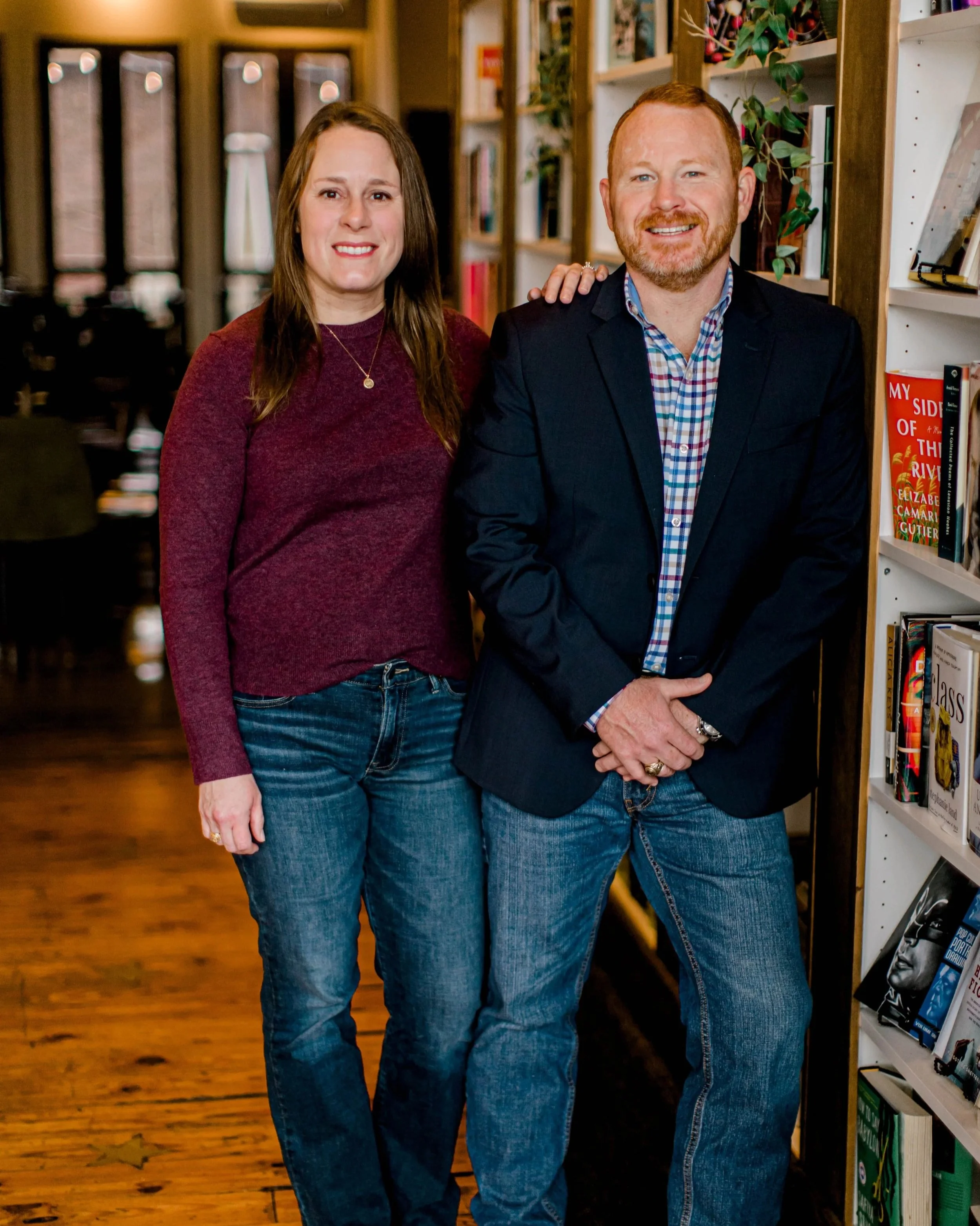 A woman with long brown hair and a man with short red hair and beard standing together in a bookstore, smiling at the camera. The woman is wearing a maroon sweater and jeans, and the man is wearing a blue blazer, a checkered shirt, and jeans. There are bookshelves filled with books behind them.