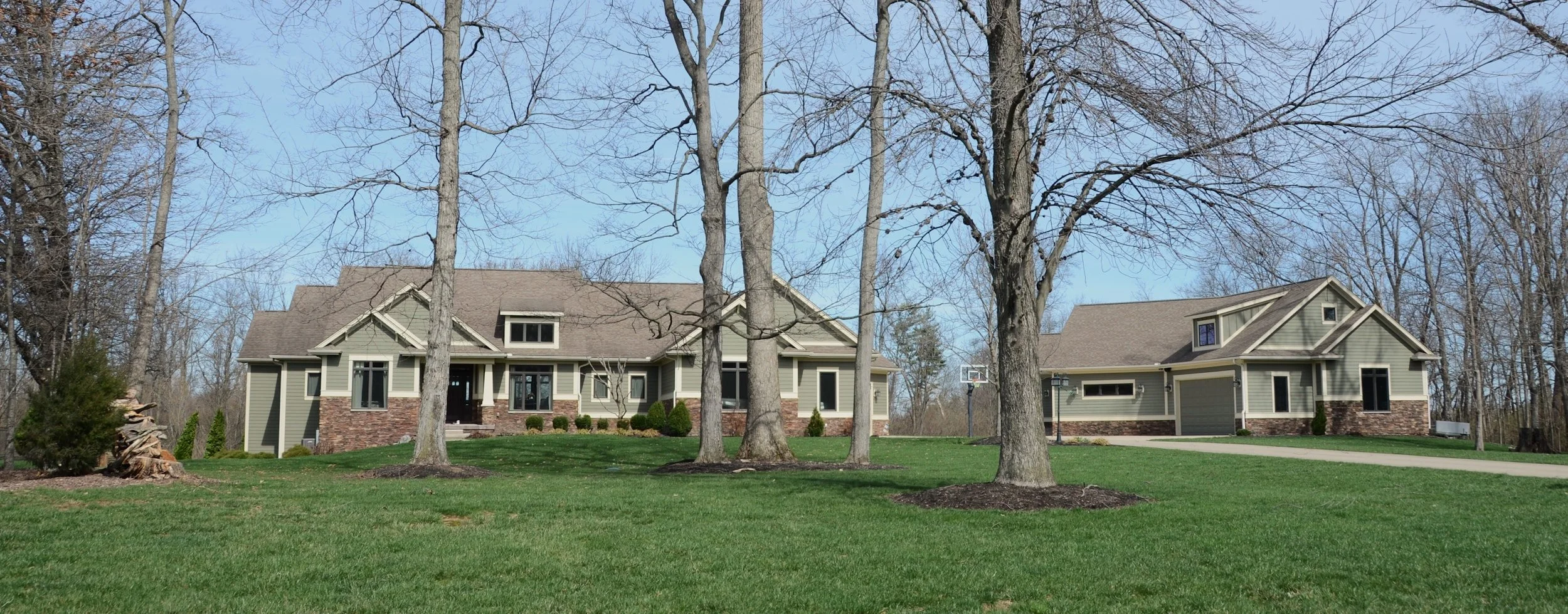 A large house with a detached garage, surrounded by a well-maintained lawn and several trees, under a clear blue sky.