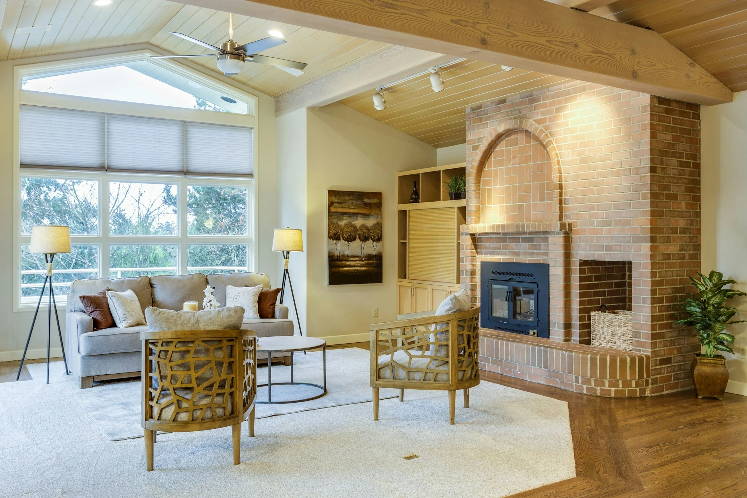 Cozy living room with beige sofa, wooden coffee table, two wooden chairs, large window with blinds, brick fireplace, and wooden ceiling with ceiling fan and track lighting.