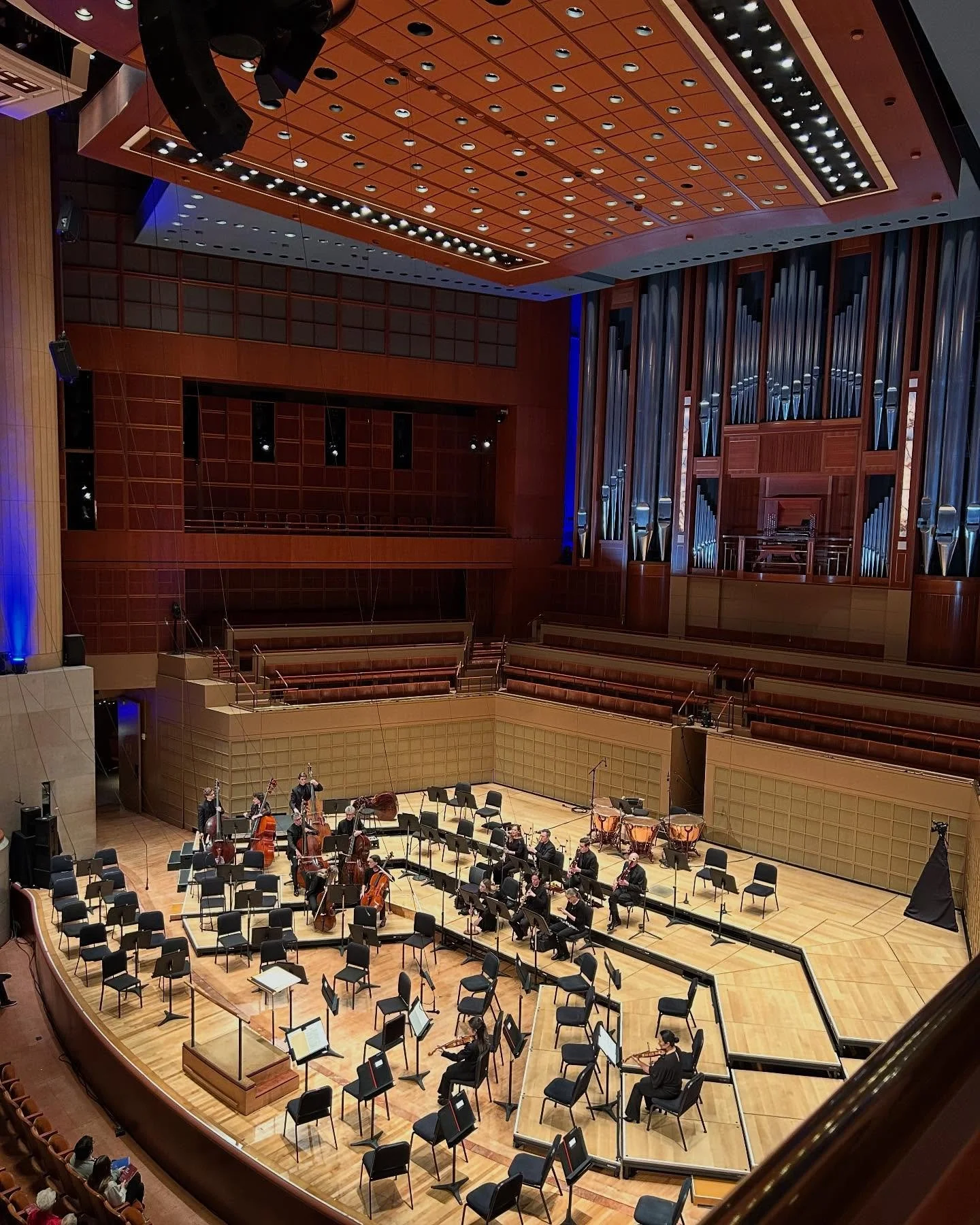 Orchestra stage in a concert hall with musicians and instruments, including violins and cellos, with empty chairs and a large organ in the back.