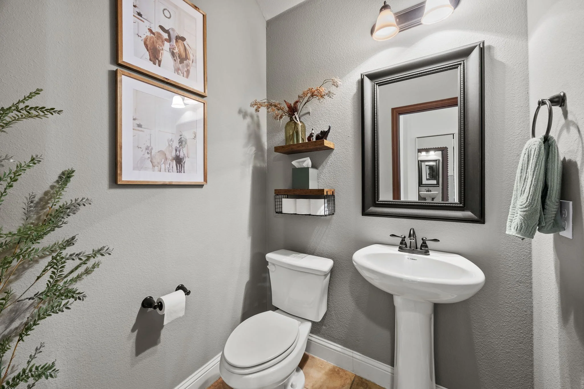 A bathroom with a toilet, a pedestal sink, and a large black framed mirror. There are two framed animal art prints on the wall, a small wall shelf with a vase, tissue box, and decorative items, a towel hanging on a ring, and a wall-mounted light fixt