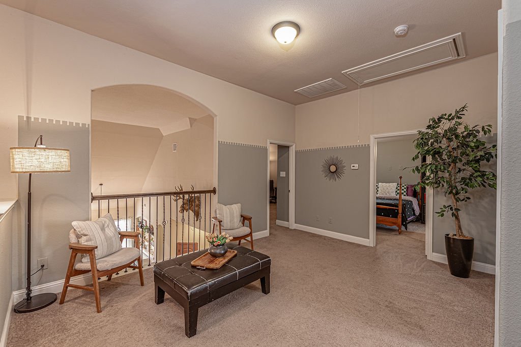 A living room with beige carpet, two armchairs, a leather ottoman, and a potted plant, with an open doorway leading to a bedroom.