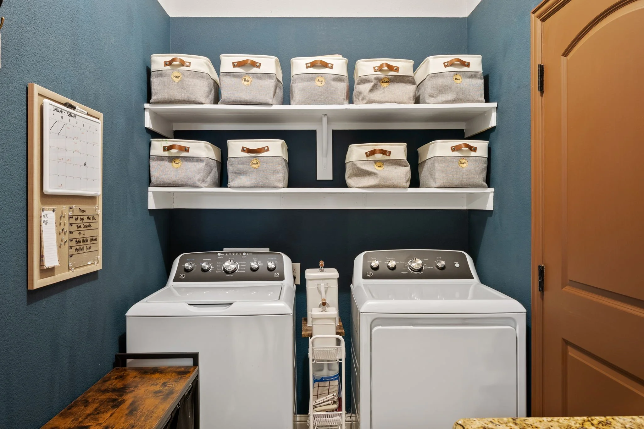 A laundry room with a matching white washer and dryer, two white shelves above with labeled storage bins, a wooden bench to the side, a small white cart between the appliances, and a wall-mounted calendar on the left side.