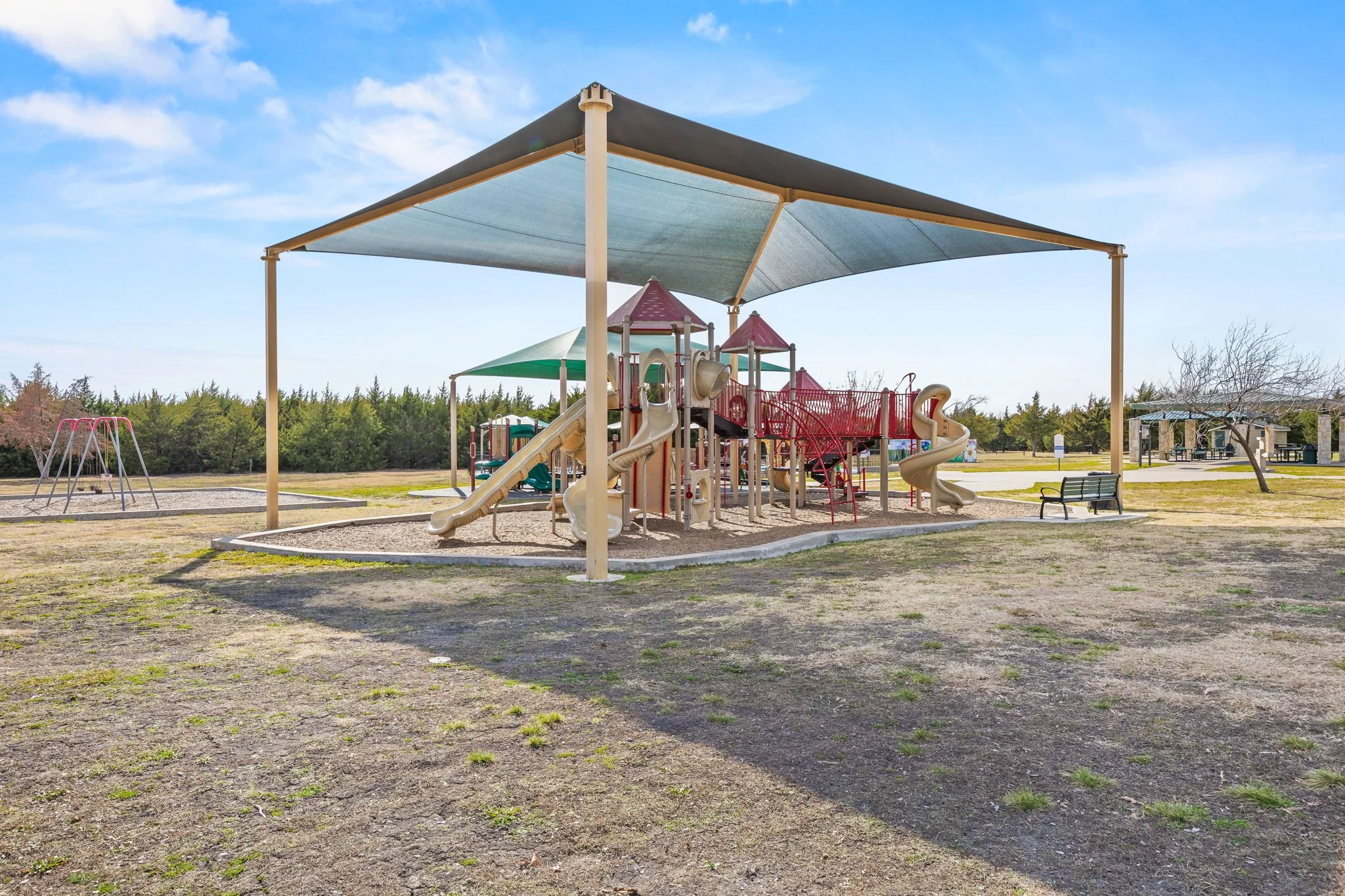 Empty playground with a large play structure featuring slides and bridges, shaded by a canopy under a blue sky on a sunny day.