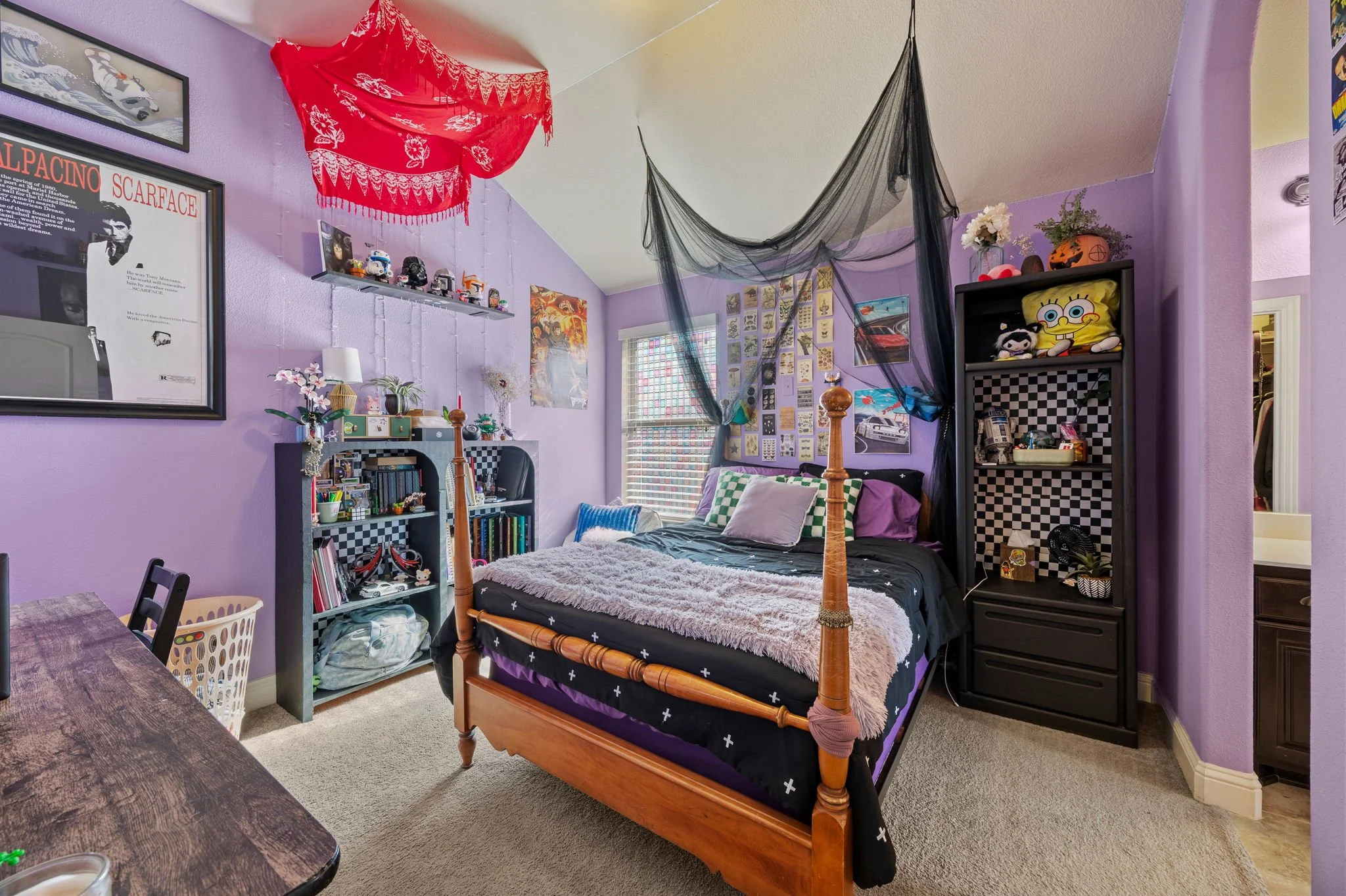 A bedroom with purple walls, a wooden bed with a black and purple bedspread, and various posters and decorations on the walls. There are black shelves with toys and plushies, a window with blinds, and hanging black and red fabric decorations from the