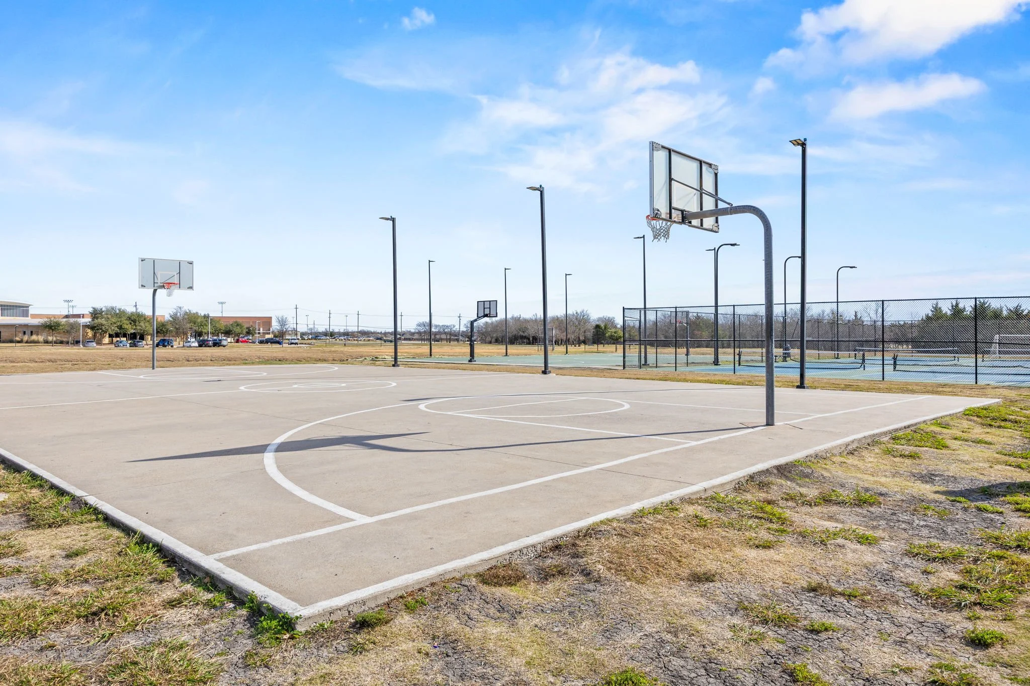 Outdoor basketball courts with multiple hoops at a park, fencing, and a clear sky.