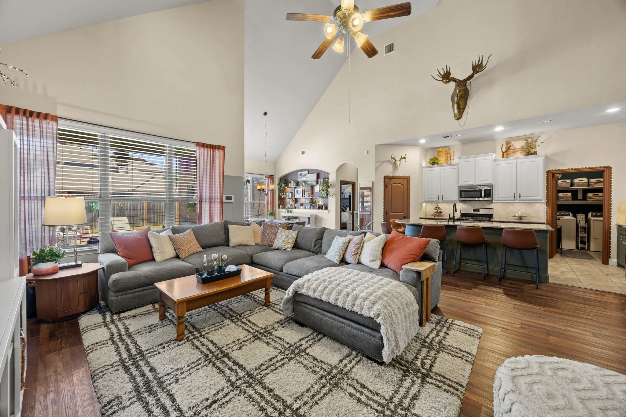 Living room with gray sectional sofa, beige and rust-colored pillows, wooden coffee table, and patterned area rug. Large window with pink curtains, coastal ceiling fan, and mounted deer head. Open kitchen with white cabinets, black countertops, and b