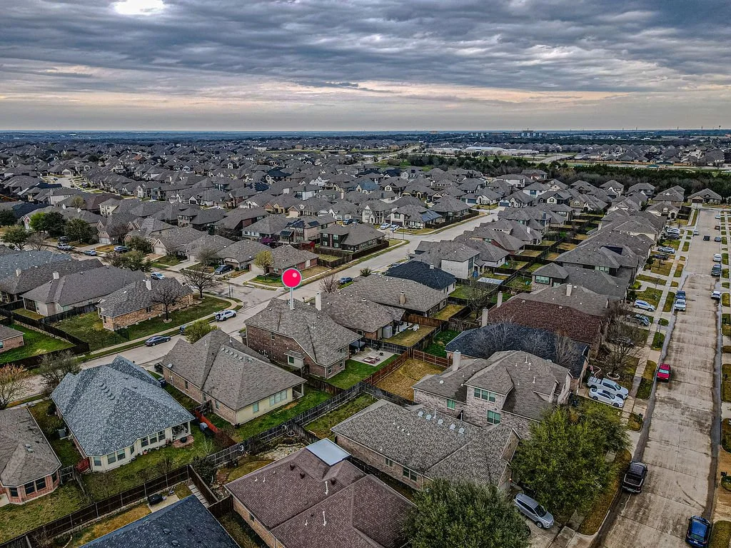 Aerial view of a suburban neighborhood with rows of single-family houses, parking lots, and tree-lined streets under a cloudy sky.