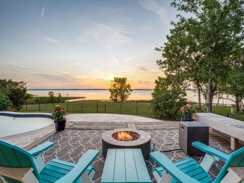 A backyard patio with a fire pit, pool, chairs, potted plants, and a view of a lake at sunset.
