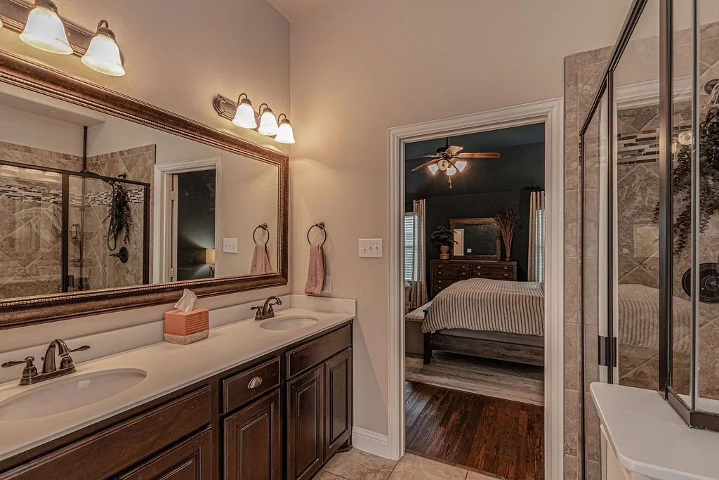 A bathroom with a double vanity, large mirror, and lighting fixture, adjacent to a bedroom with a bed, dresser, and ceiling fan.