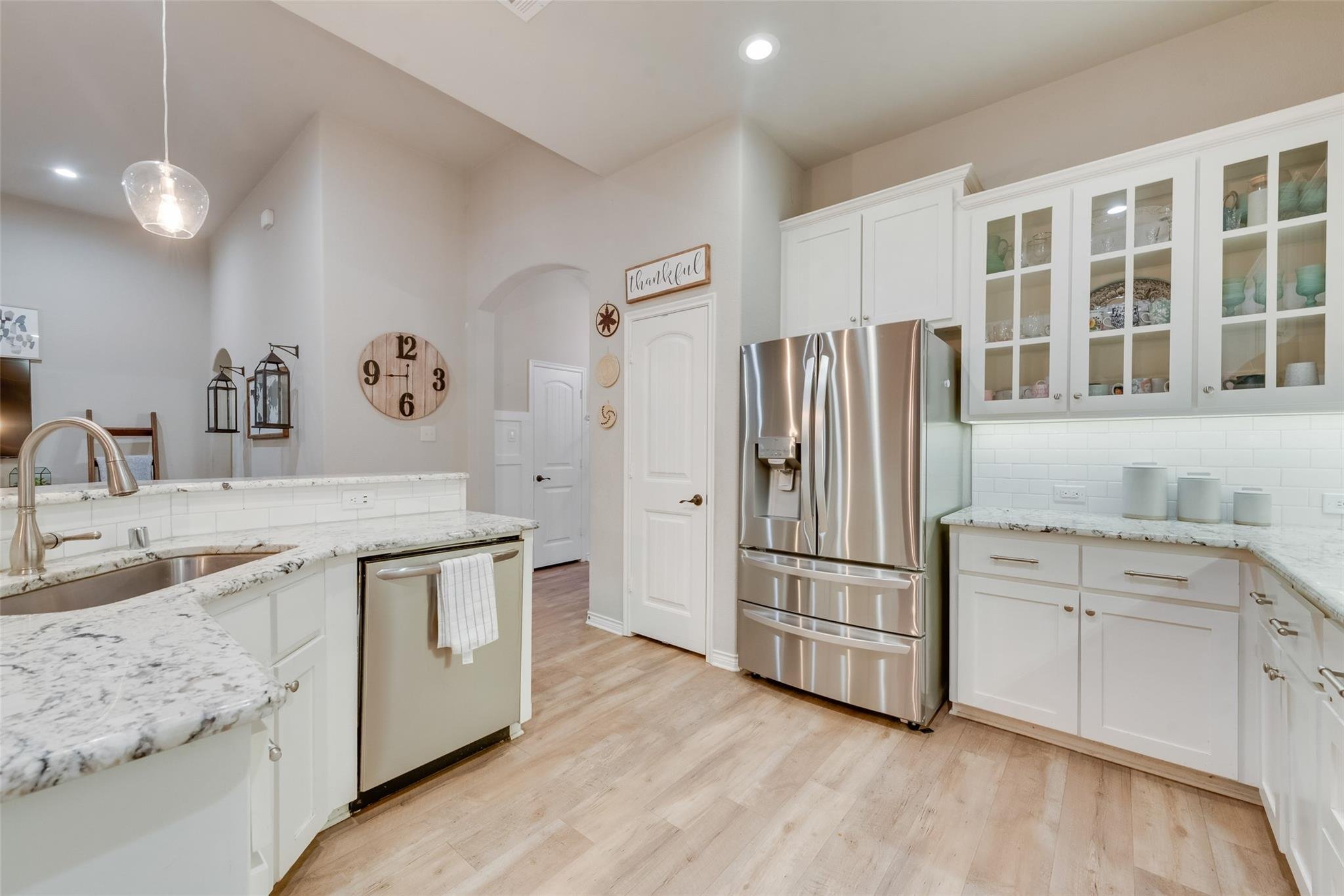 Modern kitchen with stainless steel appliances, white cabinets, marble countertops, and wooden flooring. Features a visible clock, a pendant light, and decorative accessories.