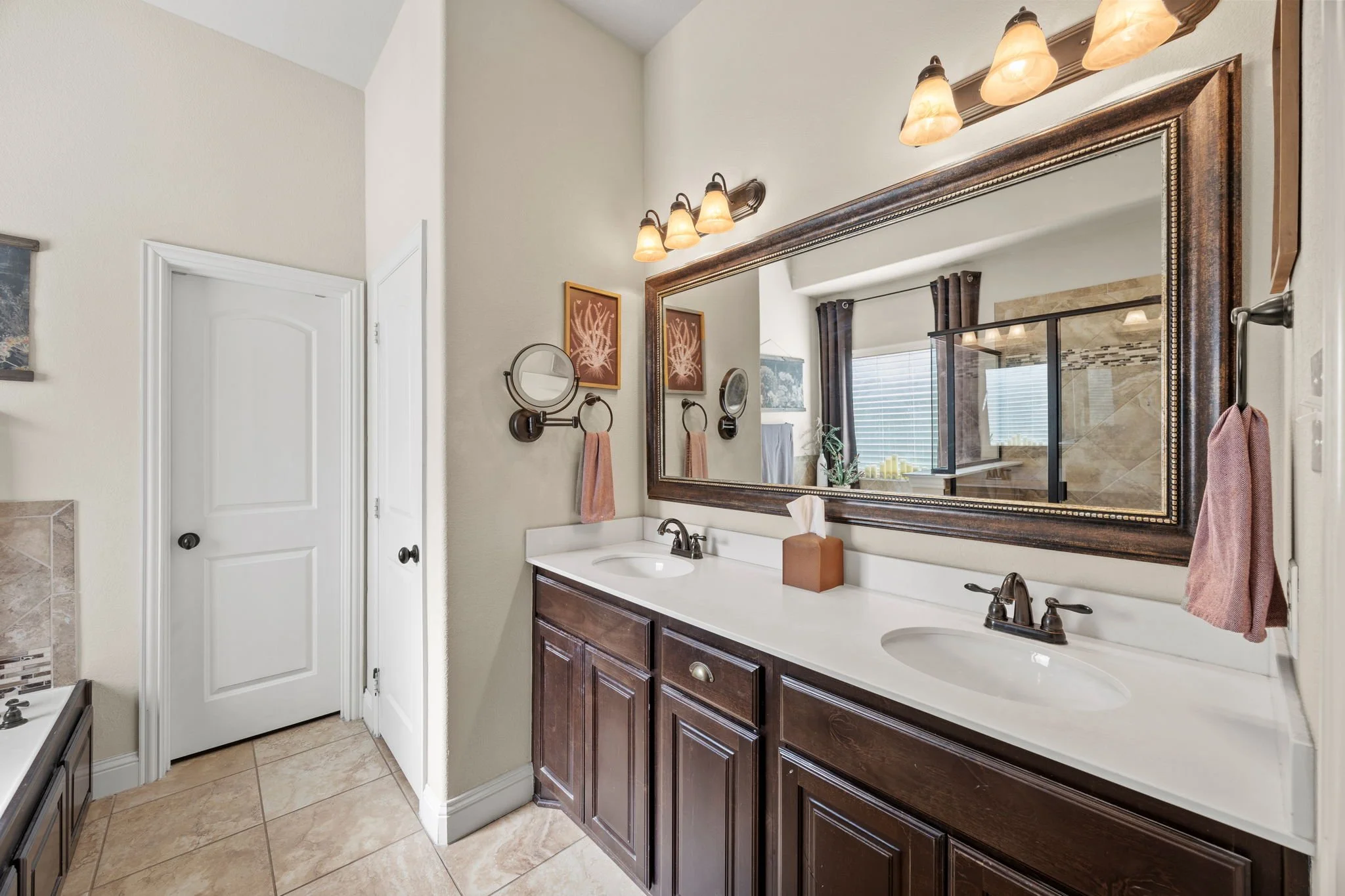 Bathroom with double sink vanity, large mirror, wall-mounted lights, towel ring with pink towel, tissue box, and decorative wall art, with a window and shower in the background.