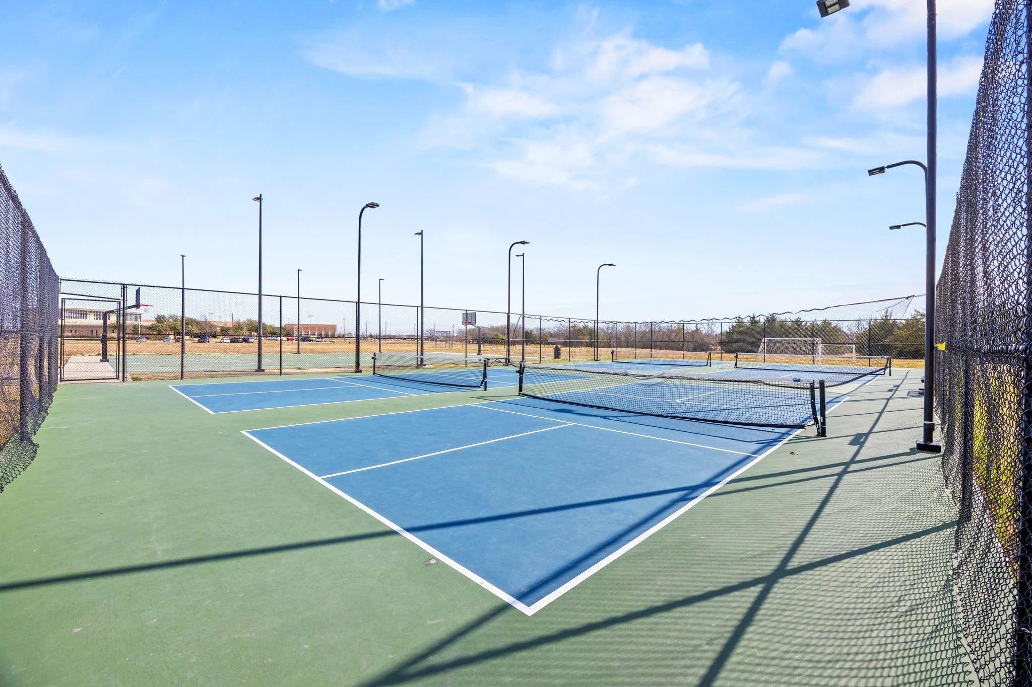 Empty outdoor tennis courts with blue and green surfaces, surrounded by black fencing, under a blue sky with some clouds, and distant trees and buildings.