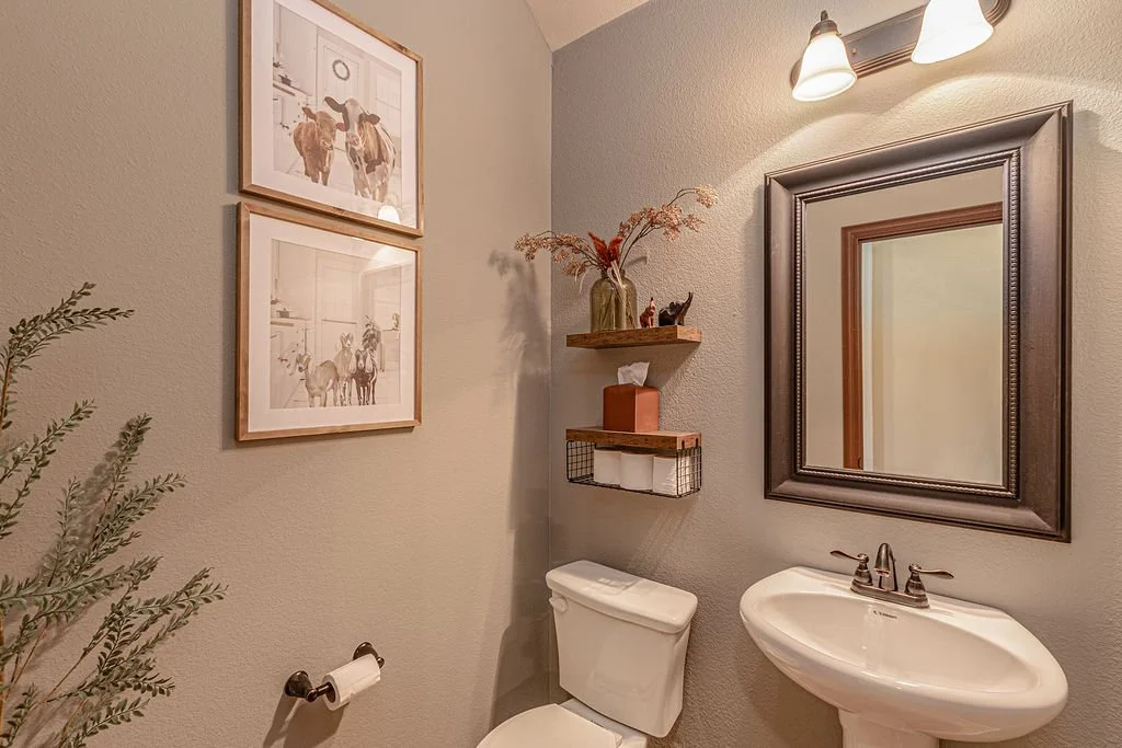 Small bathroom with a mirror, sink, toilet, wall-mounted shelves holding decor and tissue box, framed artwork, wall-mounted light fixtures, and a plant.