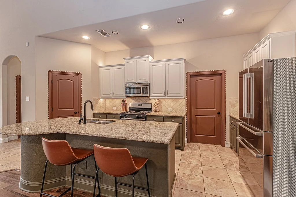 Modern kitchen with a large island, granite countertops, stainless steel appliances, and white and green cabinets. Two brown bar stools are at the island, and there are two wooden doors in the background.