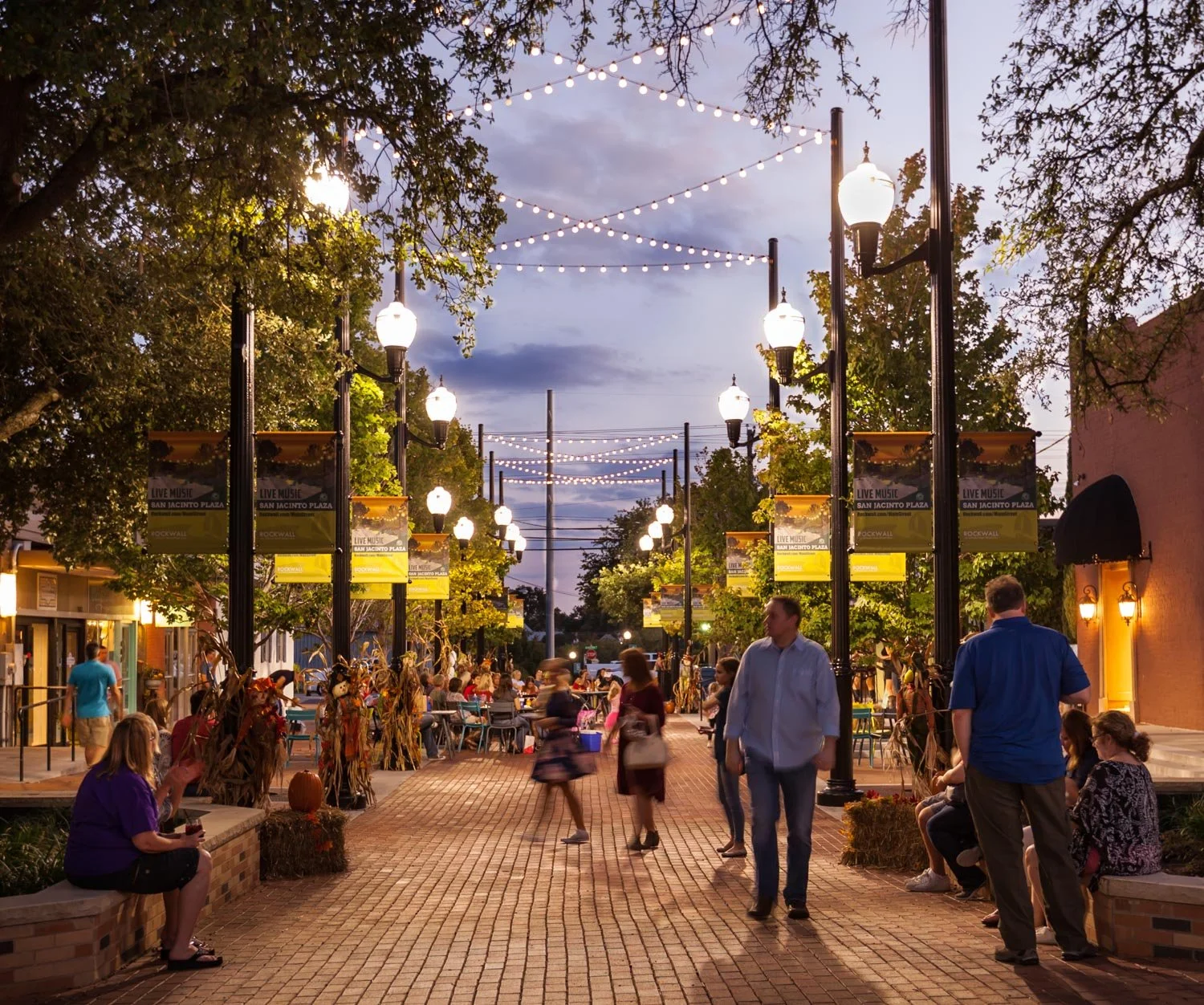 People walking and sitting on benches along a lively outdoor shopping district at dusk, with string lights and street lamps illuminating the scene.