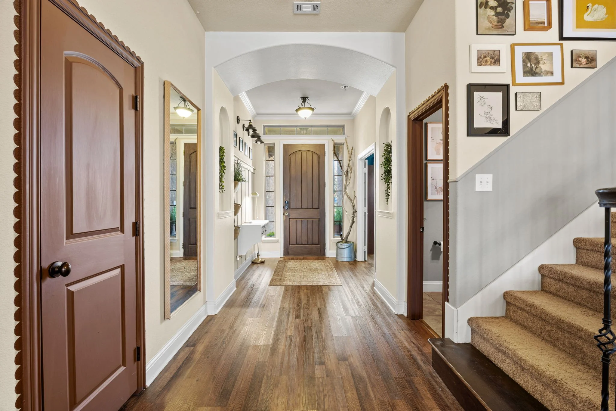 A spacious, well-lit foyer with a wooden front door, hardwood floors, and a staircase with beige carpet. Interior features include framed artwork on the wall, a mirror, a small wall-mounted sink, and decorative plants.