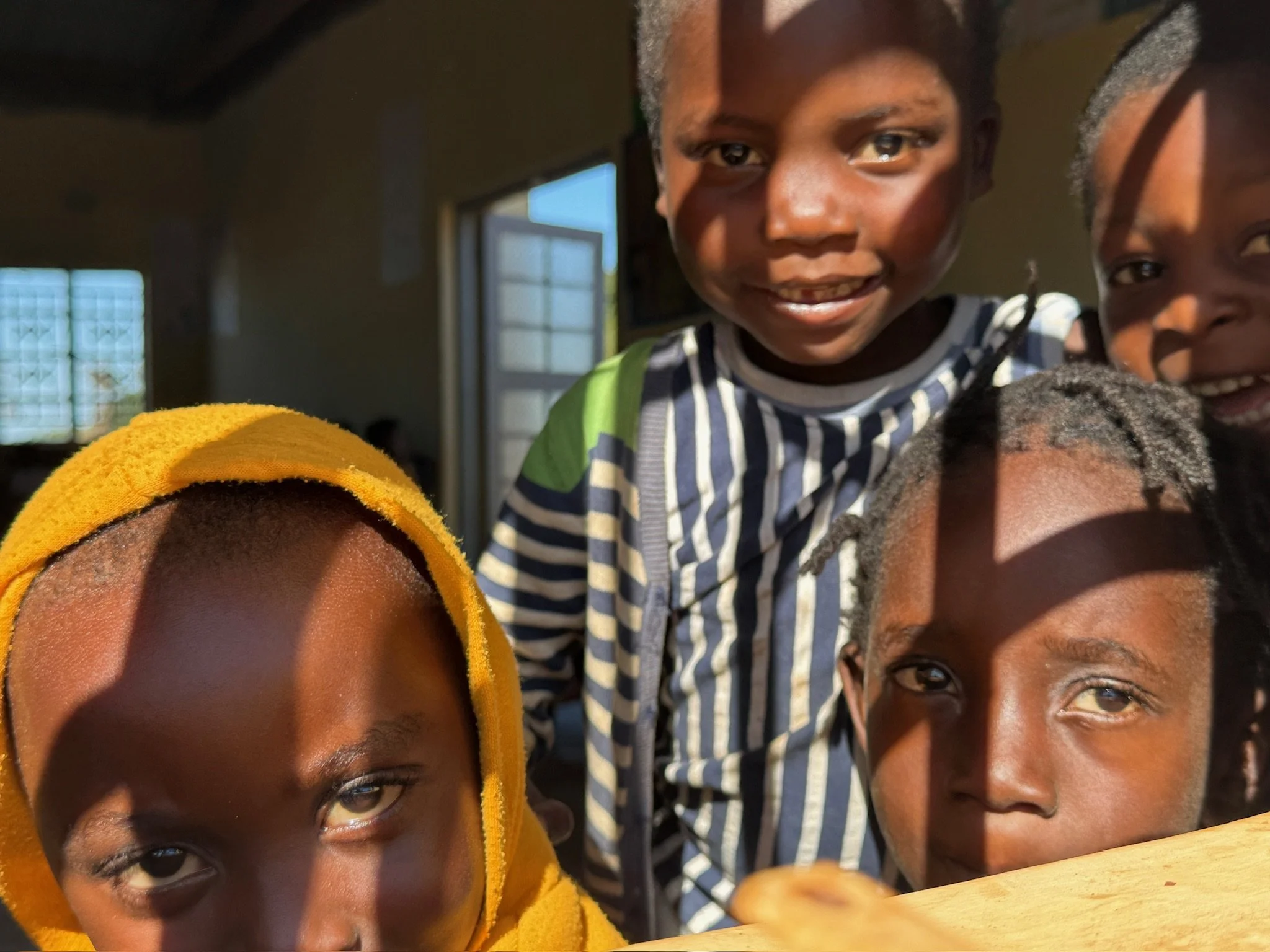 Four children with dark skin smiling and looking at the camera indoors, with sunlight casting shadows on their faces.