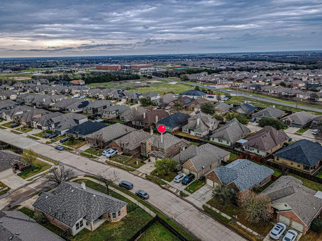 Aerial view of a suburban neighborhood showing houses, streets, and parked cars under a cloudy sky.