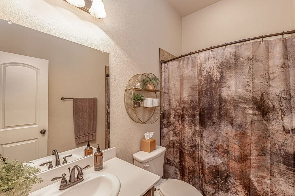 A bathroom with a white sink, a large mirror, brown towels, a tissue box, a round wall shelf with plants and toiletries, a decorative shower curtain, beige textured walls, and a white door.