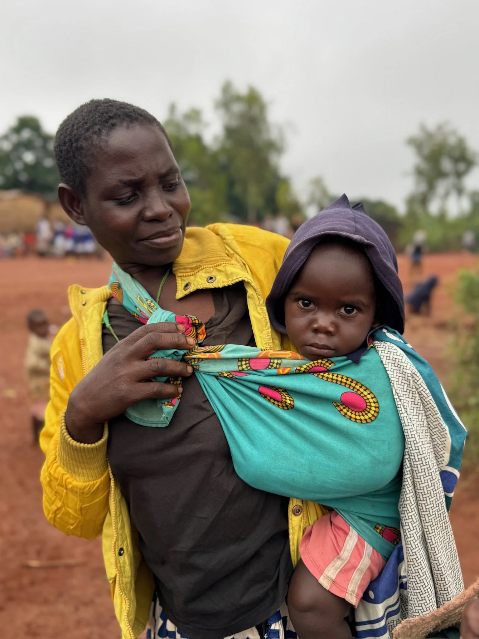 A woman in a yellow jacket holds a young child in a colorful wrap in an outdoor rural setting with trees and other children in the background.