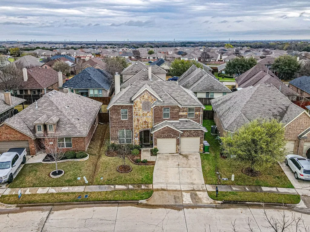 Aerial view of a suburban neighborhood with multiple houses, mostly brick with shingled roofs, front lawns, trees, and driveways, under a cloudy sky.