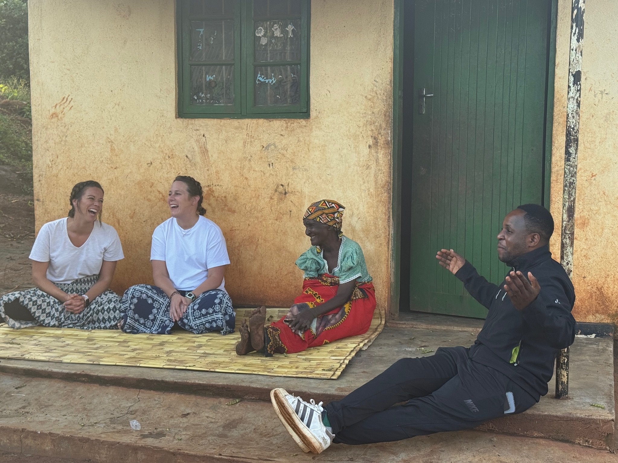 Four people sitting outside a yellow building with a green door and window, laughing and engaging in conversation. The group includes three women and one man, dressed casually, with a man sitting on the ground with his back against the building, gest