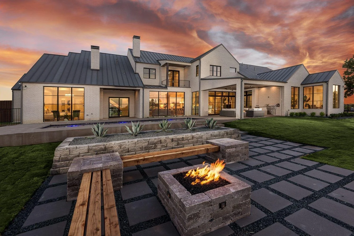 A modern house with large glass windows, lit from inside, at sunset. The backyard features a fire pit, outdoor seating, a pool, and well-maintained lawn.