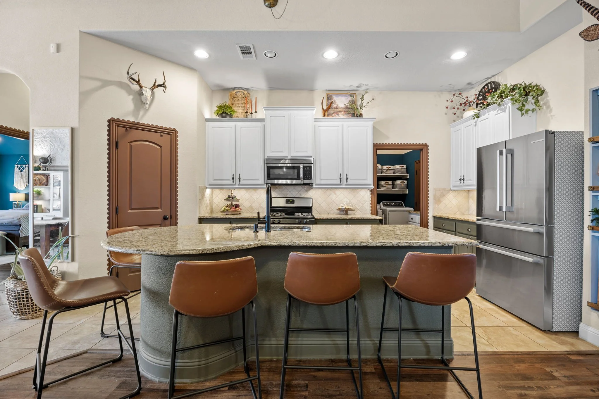 Modern kitchen with white cabinets, stainless steel appliances including a refrigerator and microwave, granite countertops, and a large kitchen island with brown leather chairs. Decor includes a mounted deer skull, greenery, and a view into a laundry