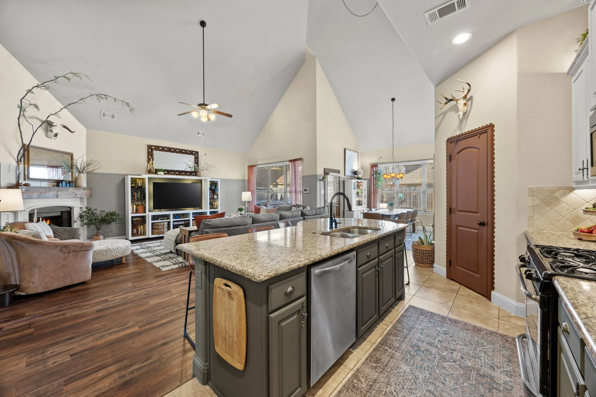 Open-concept living room and kitchen with vaulted ceiling, granite island, fireplace, TV, and large windows, decorated with plants and mounted animal skulls.
