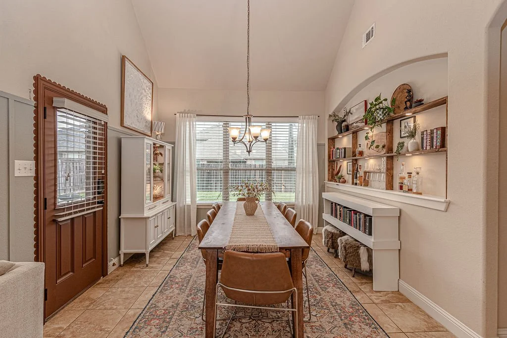 Dining room with a wooden table, six chairs, a white sideboard, white curtains, a chandelier, built-in shelves with various decorative items and books, and a window showing a backyard.
