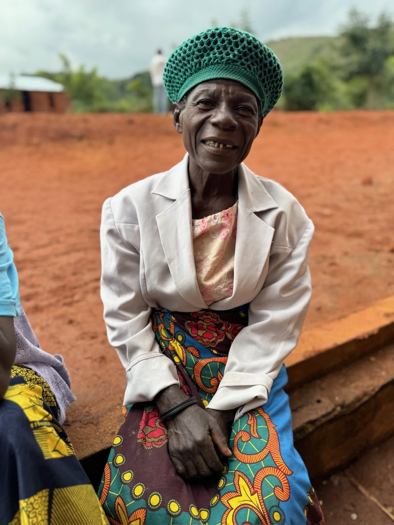 An elderly African woman wearing a green hat, white blouse, and colorful patterned skirt, sitting outdoors on a reddish-brown ground with green hills and trees in the background.