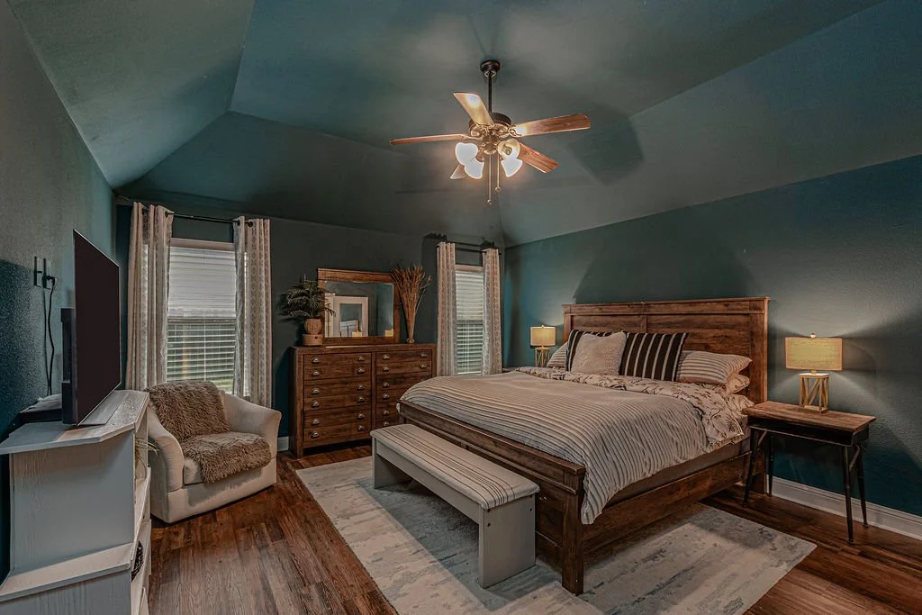 A cozy bedroom with a large wooden bed, striped bedding, nightstands with lamps, a wooden dresser with a mirror, a cream armchair with a furry cushion, and three windows with curtains. Has a ceiling fan and hardwood floors.