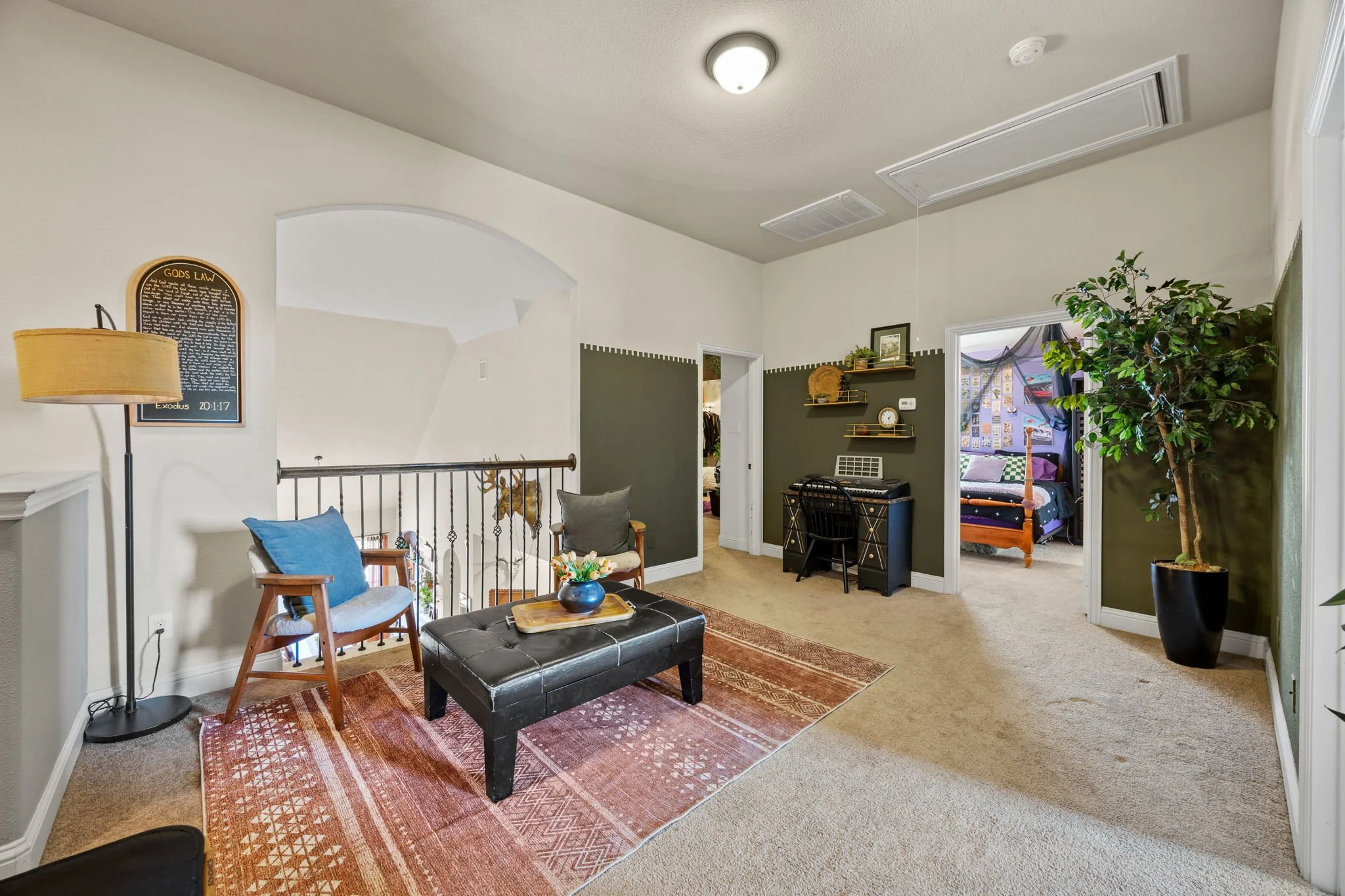 Living room with beige carpet, green and white walls, a black ottoman, a wooden chair with a blue cushion, a floor lamp, a small desk with a keyboard, shelves, and a large potted plant. An open doorway leads to a bedroom with a canopy bed.
