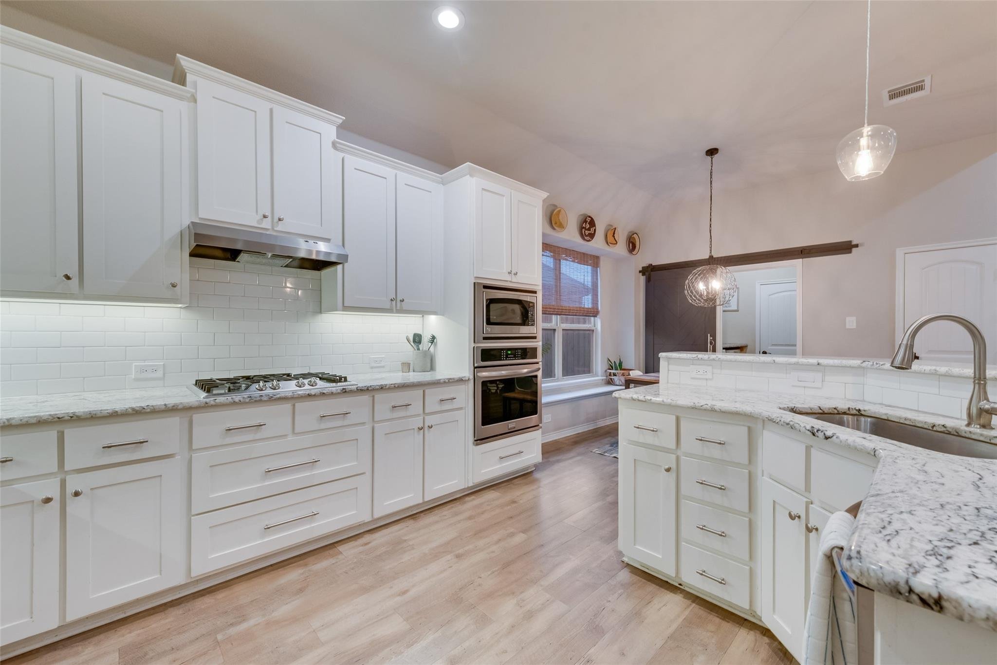 Modern kitchen with white cabinets, marble countertops, a stainless steel oven, and a stove. There's a dining area with a window and pendant light, wood flooring, and a barn-style sliding door.